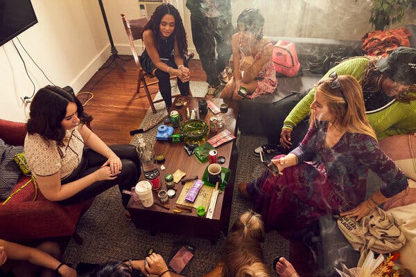 A group of people sit in a smoky living room around a coffee table covered in cannabis paraphernalia, including buds and bongs.